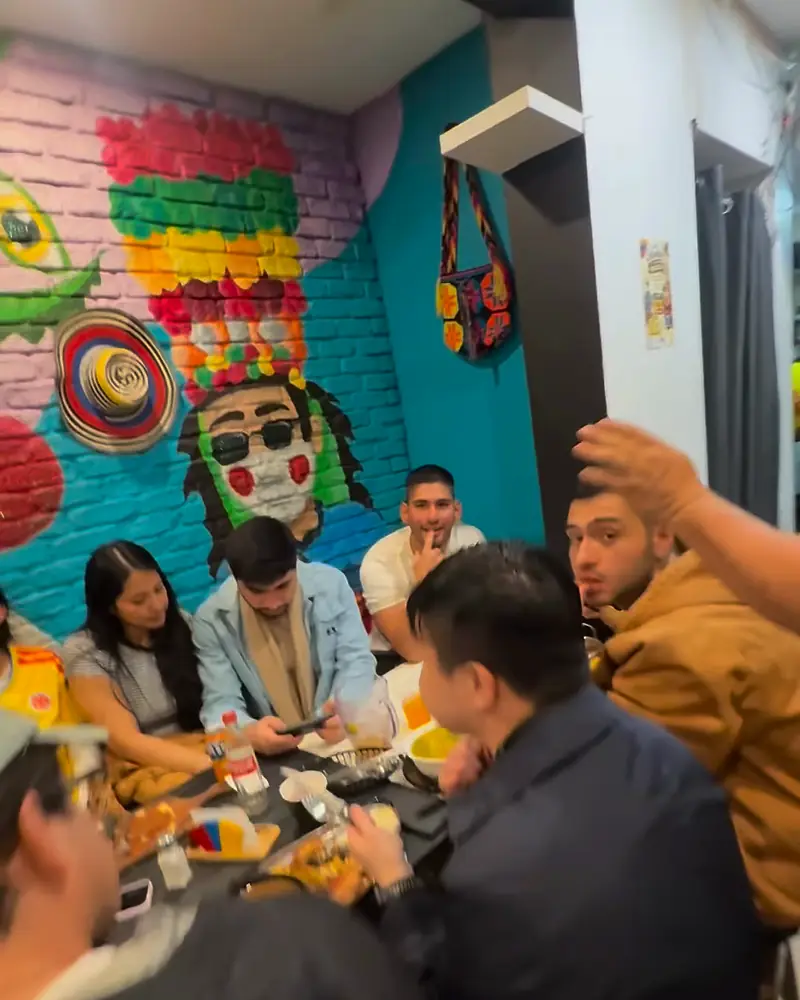 Diners at a long table under a painted Carnaval de Barranquilla mask mural on a teal and pink brick wall