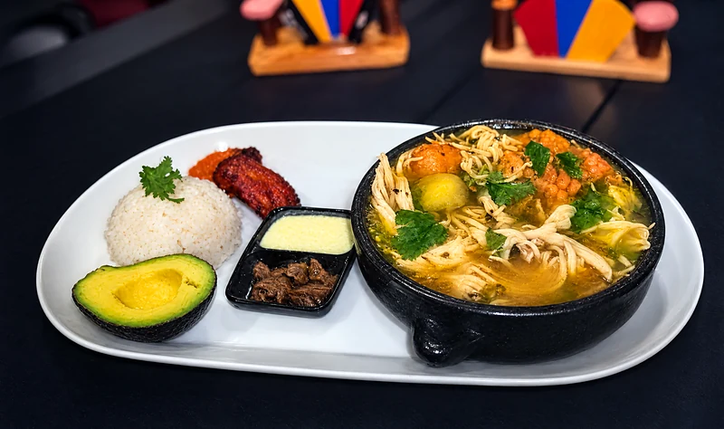 Ajiaco in a black clay pot with rice, avocado, fried plantain, cream and capers on the side