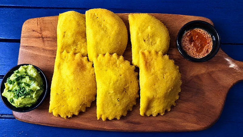 Six golden empanadas on a wooden board with guacamole and a small bowl of sauce