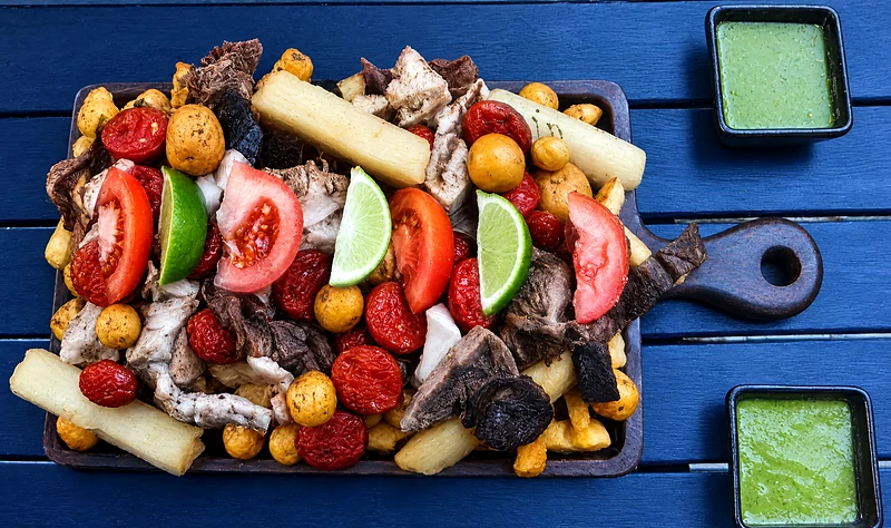 Picada platter: chicharron, chorizo, black pudding, potatoes, cassava, tomato and lime on a blue table