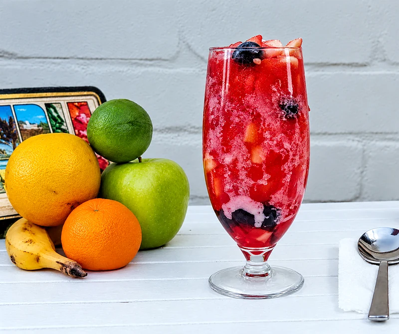 A tall glass of salpicon, chopped fruit in red juice, beside a lemon, lime, apple, orange and banana