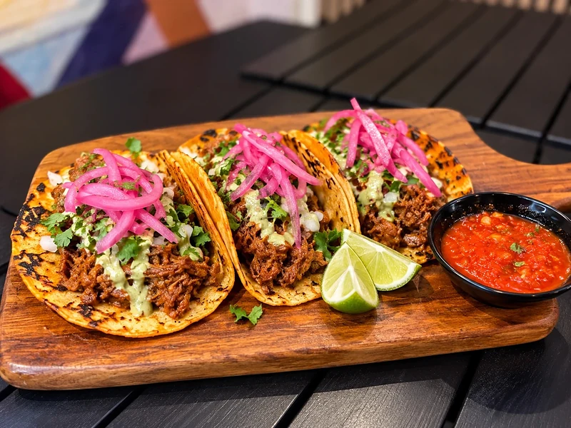 Three tacos with pulled meat, pickled onion, coriander and a green sauce on a wooden board, with lime and salsa